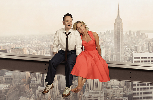 Joe Stilgoe and Liza Pulman, dressed in 1920s style clothing, sit on a steel beam above New York City