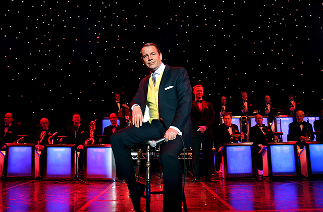 Stephen Triffit on stage as Frank Sinatra in a sharp black suit with yellow waistcoat, and a large band behind him. Above, lights emulate stars in the night sky.