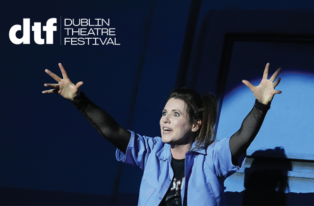 Naomi Louisa O'Connell wearing a short-sleeve blue shirt, eyes wide, mouth open, with her arms raised, against a dark blue background. There is a Dublin Theatre Festival logo top left.