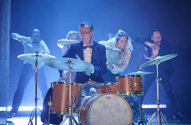 Conor Guilfoyle drumming in a tuxedo, with dancers moving behind him.