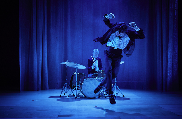 A heavily blue-filtered image of Alex O'Neill in a tuxedo mid-jump, with his hands above his head. On the stage behind him, Conor Guilfoyle, also in a tux, plays a drum kit.