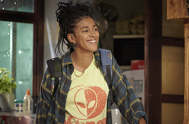 Fanta Barrie smiling, wearing a yellow t-shirt with a check overshirt. She has a backpack on.