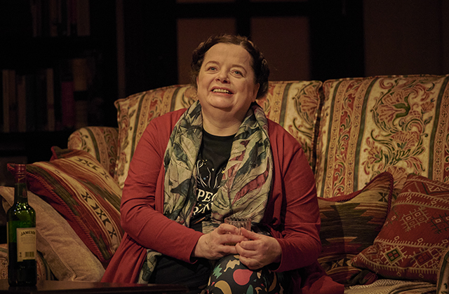 Helen Norton, smiling, sitting on a patterned couch. She is wearing a red top with a floral scarf.