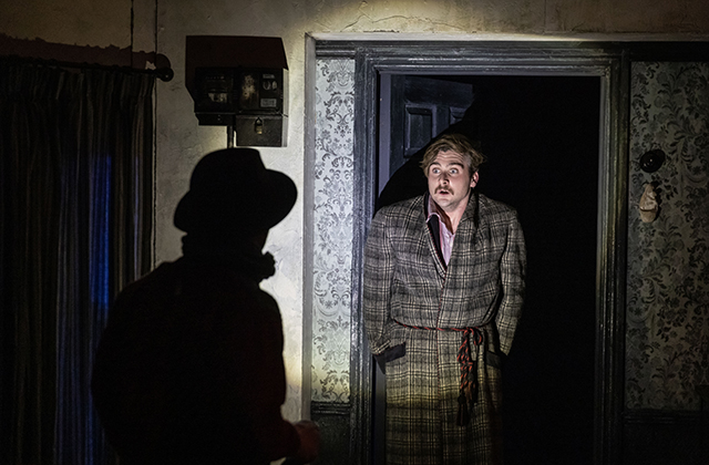 Liam Heslin, in a dark room, caught in the glow of a torch shone by a silhouette in the foreground. He is wearing a housecoat.