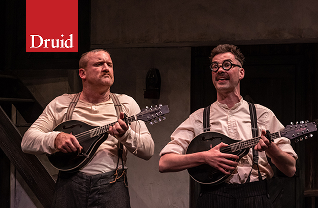 Rory Nolan and Marty Rea, both with playful expressions and playing mandolins, wearing white shirts and suspenders.