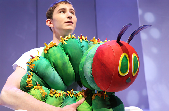 Close-up shot of the Very Hungry Caterpillar puppet, appearing as he does in the iconic illustration. A puppet performer holds him.