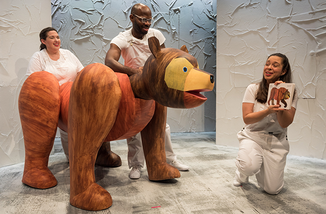 A large puppet of Brown Bear. He is puppeted by two performers, and looking at another performer who holds open the picture book Brown Bear, Brown Bear in a very meta tableau.