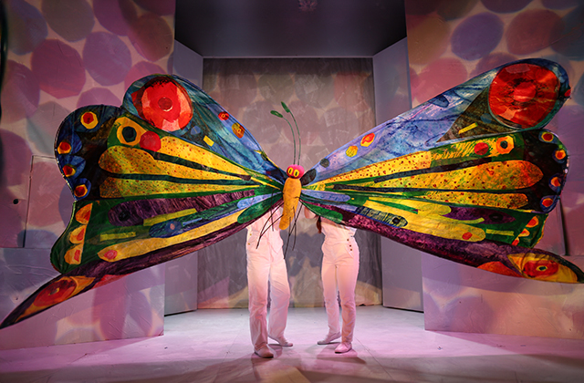 A giant puppet butterfly, its wings many meters wide in kaleidoscopic colours.