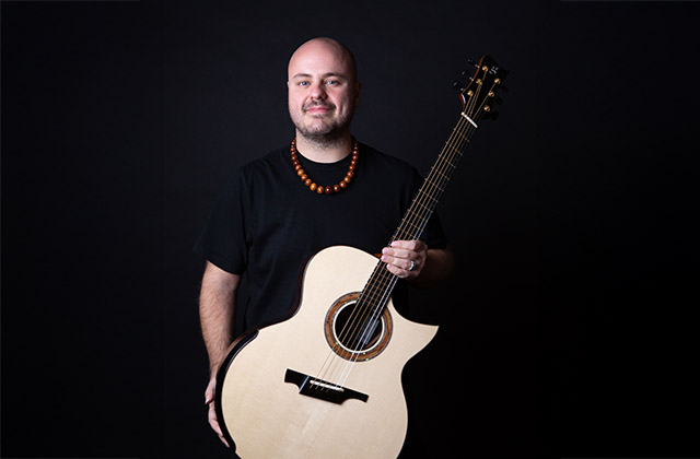 Andy McKee stands with his guitar in his hands held across his front against a black background, he is looking towards the camera smiling, he is wearing a black t-shirt.