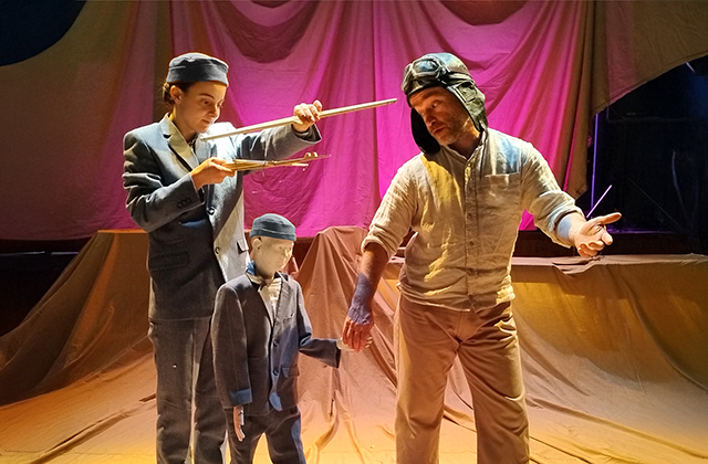 Two actors are on stage together, one has a puppet on a string that is dressed in the same costume as the puppeteer, the other is a man who is wearing an old style pilots hat, there is some red and yellow material draped behind them on the stage. The pilot is staring down to look at the puppet.