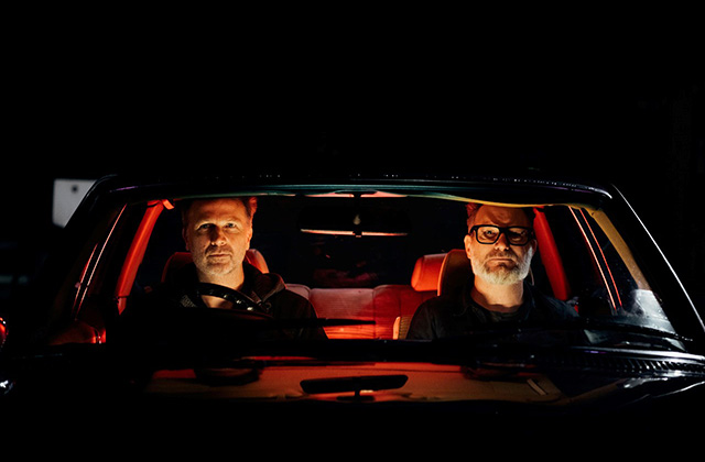 Paul Noonan and Brian Crosby are sitting together in the front seats of a car looking through the front windshield straight to the camera. The photo is dark and it appears to be night time, there is a red hue coming off the cars interior and from Paul and Brian. Photo by Johnny Malin.