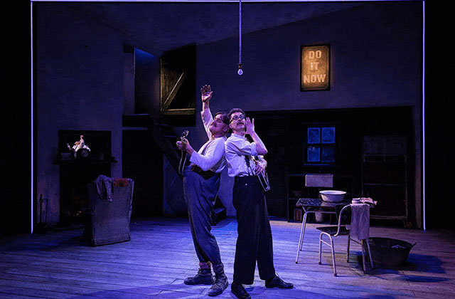 Rory Nolan and Marty Rea are standing together back to back on stage holding their mandolins under blue lighting. They are casting their arms and gaze upwards and appear to be singing a song.