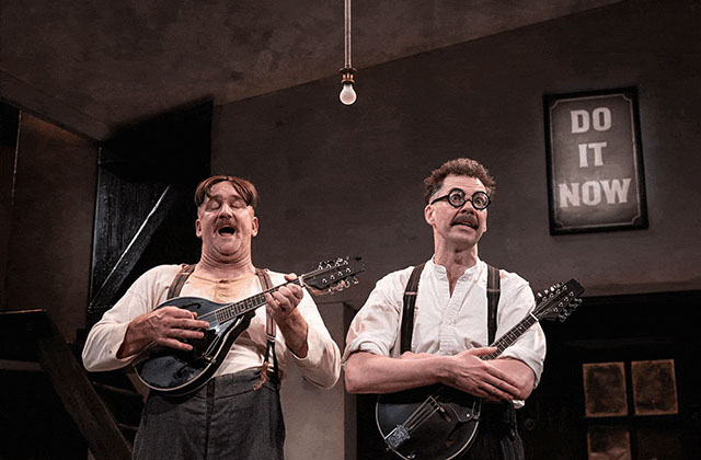 Rory Nolan and Marty Rea are standing together on stage holding mandolins. They are wearing white shirts with dark suspenders and appear to be singing a song.